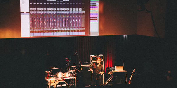 David Carr stands on stage in front of a drum kit while a screen recording of a mixing software is projected on the wall behind him